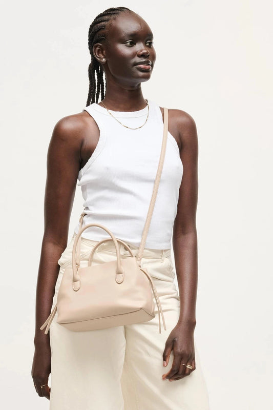 A woman wearing a white tank top and cream pants carries a light beige leather crossbody handbag with dual top handles and a thin shoulder strap against a plain off-white studio background.