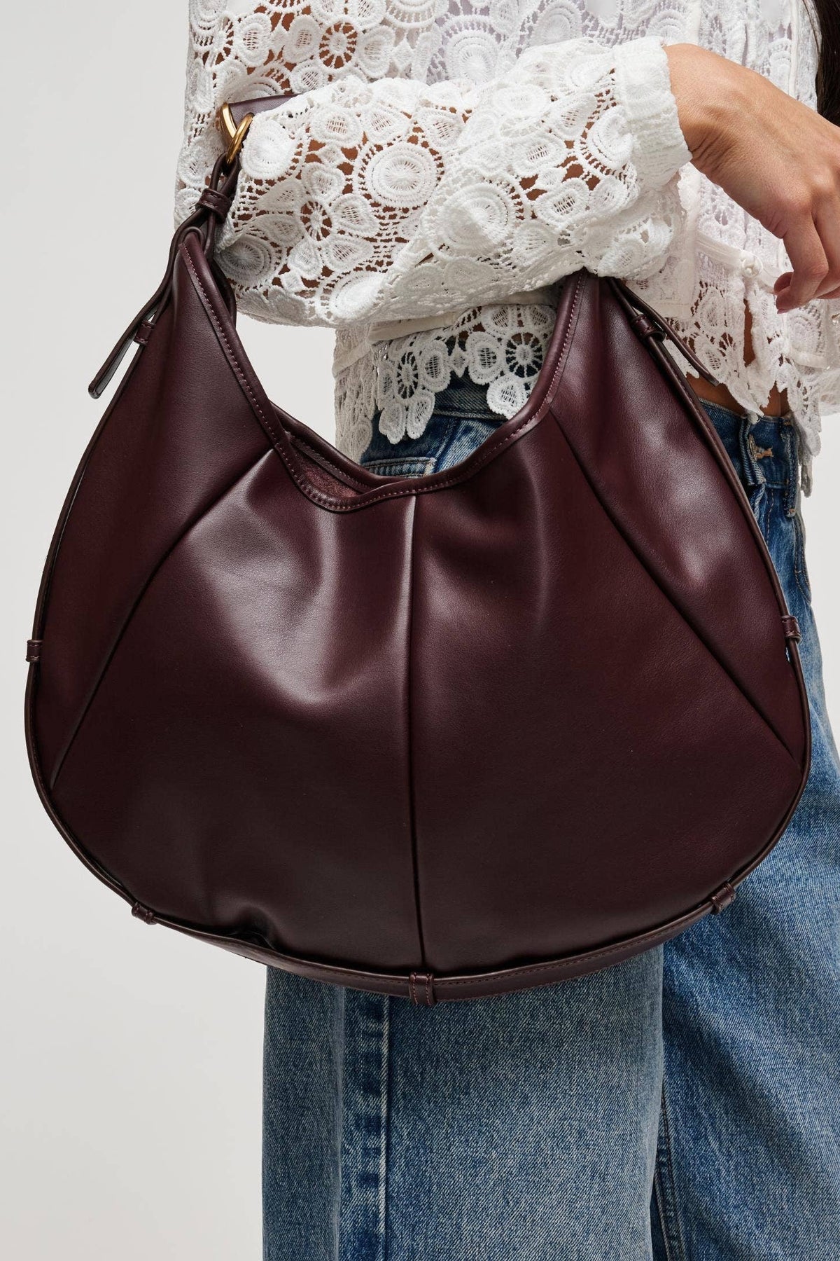 A person wearing a white lace blouse and blue jeans carries a large crescent-shaped hobo shoulder bag in dark burgundy leather against a plain light gray background.