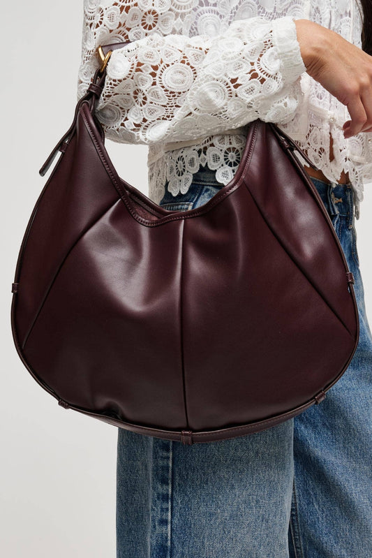 A person wearing a white lace blouse and blue jeans carries a large crescent-shaped hobo shoulder bag in dark burgundy leather against a plain light gray background.