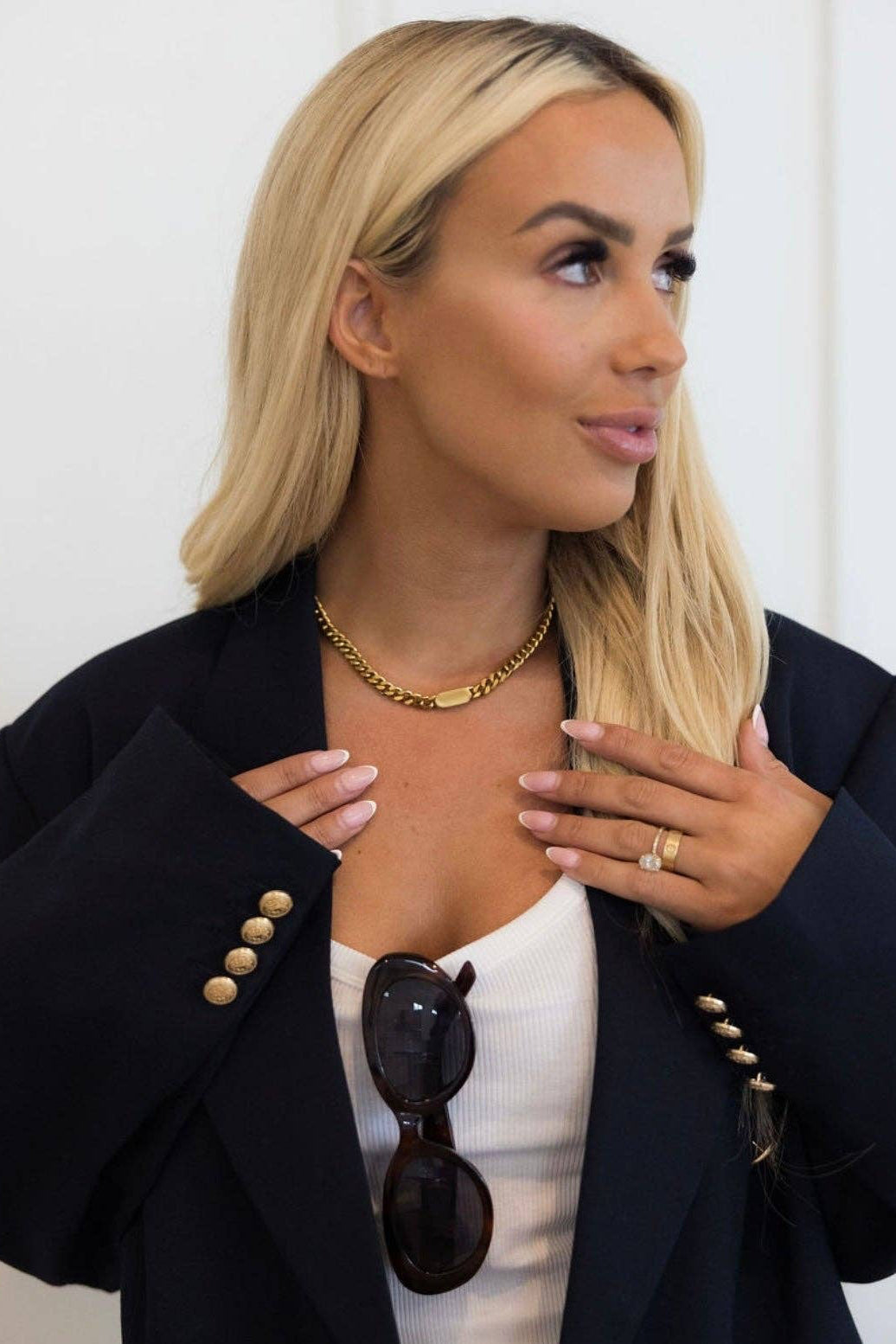 Woman wearing a black blazer with gold buttons, white top, and sunglasses on a white background