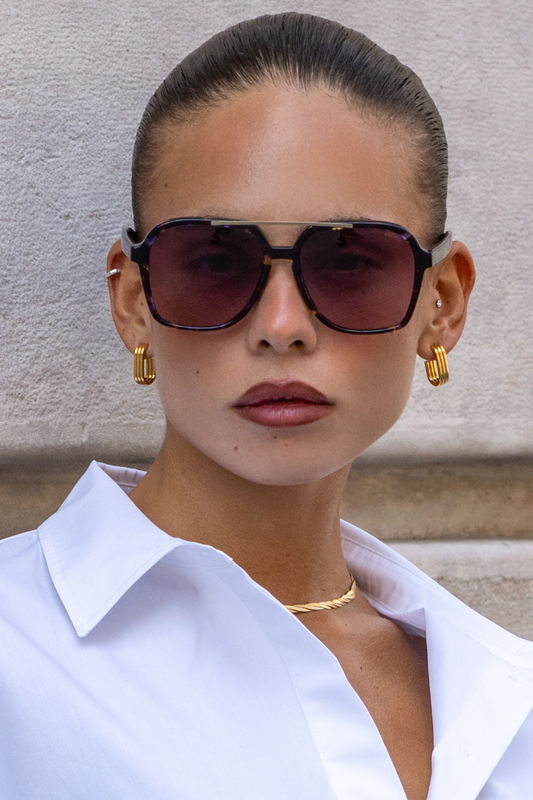 Woman wearing sunglasses and a white shirt against a stone wall.