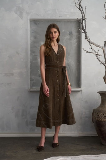 A woman models a sleeveless brown corduroy midi dress with a button-front bodice, matching tie belt, and contrast stitching, set against a textured gray wall with a decorative vase and branch.