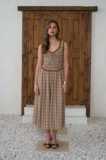A woman modeling a sleeveless, V-neck, knee-length dress with a brown and beige plaid pattern, standing in front of a rustic wooden door.