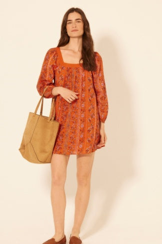 Woman wearing an orange patterned dress holding a tan bag against a beige background