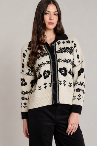 A woman modeling a cream-colored knit cardigan with black floral patterns and black trim, paired with black pants, against a solid light gray studio background.