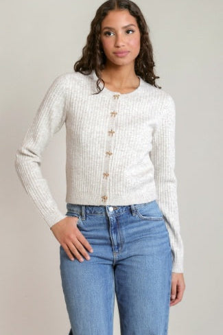 A woman modeling a cream ribbed knit cardigan with gold-tone buttons, paired with blue jeans, against a neutral light gray background.