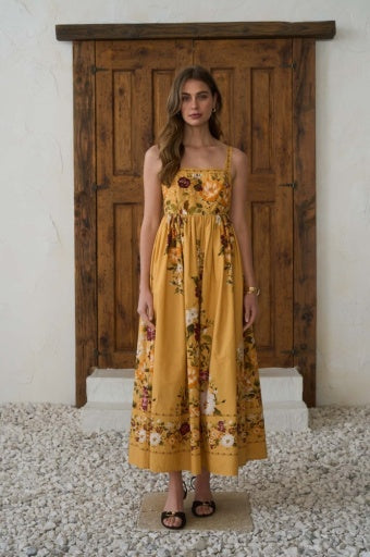 A woman stands in front of a rustic wooden door wearing a long yellow floral maxi dress with thin straps and a smocked bodice, paired with black sandals.