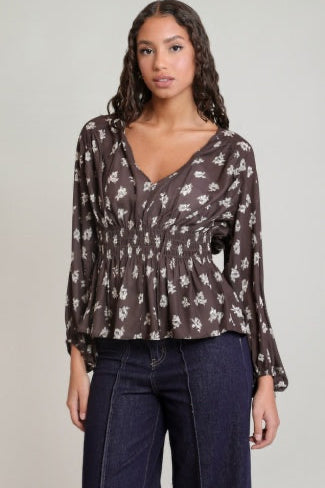 A woman modeling a dark brown long-sleeve blouse with a white floral print and a gathered waist, paired with dark blue jeans, against a plain light gray background.