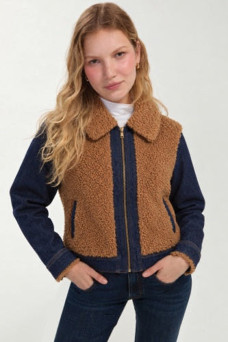 A woman modeling a dark blue denim jacket with a brown sherpa fleece lining, featuring a front zipper, a large collar, and zippered hand pockets, paired with dark blue jeans and a white t-shirt.