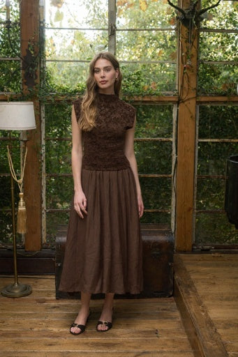 A woman stands in a rustic indoor setting with large windows and wooden floors, wearing a sleeveless brown lace top and matching brown pleated midi skirt.