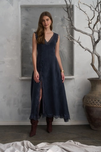 A woman modeling a sleeveless dark navy maxi dress in a rustic indoor setting with a textured gray wall, a large ceramic pot, and dried branches.