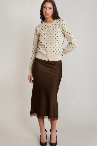 A woman modeling a cream long-sleeve knit sweater with black polka dots and a black satin midi skirt, paired with black pointed-toe heels, against a light gray studio background.