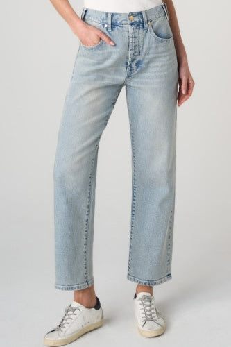 A person wearing light-wash blue straight-leg jeans with a white top and white sneakers, standing against a light gray studio background.