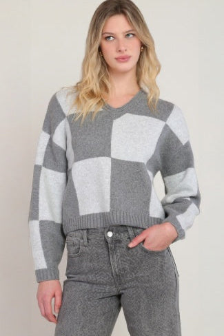 A woman wearing a gray and white checkered knit sweater with a V-neck and long sleeves, paired with gray jeans, against a plain white background.