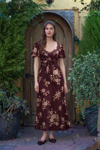 A woman stands in front of a rustic wooden door surrounded by green plants, wearing a dark brown midi dress with a beige floral pattern and short flutter sleeves.