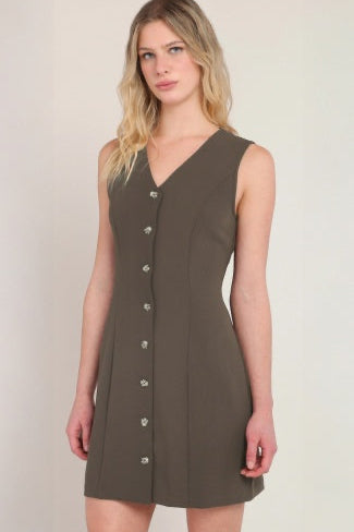 A woman modeling a sleeveless V-neck mini dress in a dark olive brown color, featuring a front button-down detail, set against a plain light beige background.