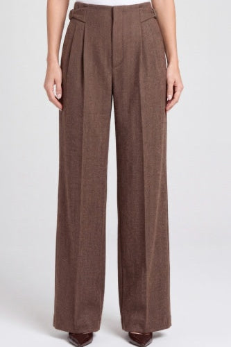 A model wearing high-waisted, wide-leg brown trousers with pleated detailing down the front, paired with dark brown pointed-toe heels against a light gray studio background.