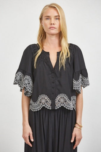 A blonde woman modeling a black short-sleeve blouse with white embroidered lace trim at the cuffs and hem, paired with a matching black skirt, against a light gray studio background.