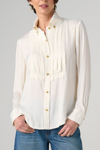 A woman wearing a cream-colored long-sleeve blouse with gold buttons and pleated detailing on the chest, paired with blue jeans, against a light gray studio background.