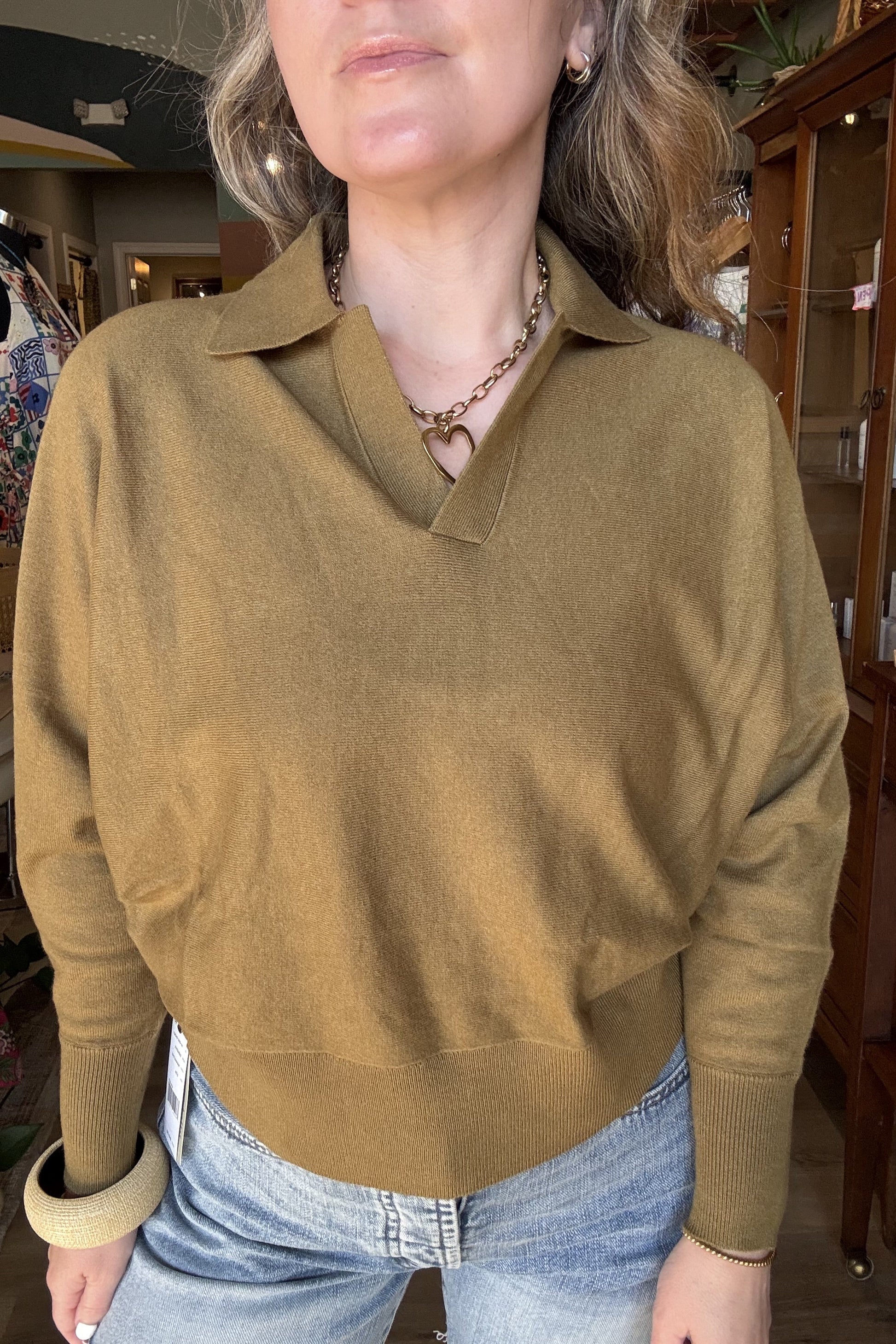 A woman wearing a camel-colored knit polo sweater with a collar and long sleeves, paired with light blue jeans, standing inside a retail store.