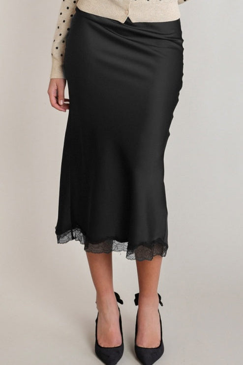 A black satin midi skirt with a sheer lace hem, styled with a cream polka-dot top and black pointed-toe heels against a light gray studio background.